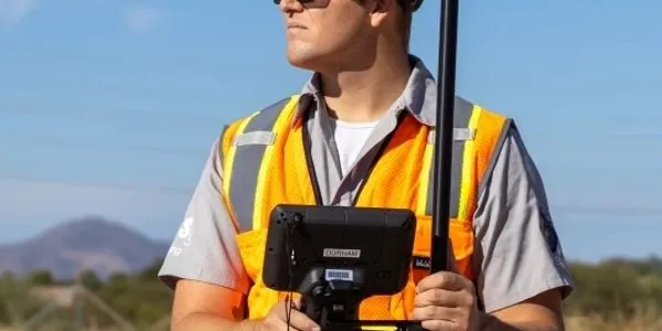 A male surveyor in a bright orange safety vest holds a GPS device, standing outdoors with mountains in the background