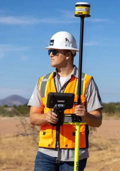 A surveyor stands outdoors in a hard hat and safety vest, holding a GPS device and pole against a clear blue sky